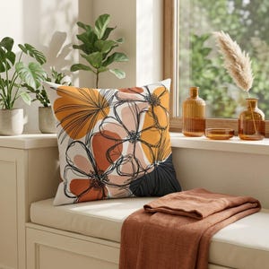 Könnte beinhalten: Ein quadratisches Zierkissen mit einem floralen Design in Orange-, Schwarz- und Pfirsichtönen. Das Kissen liegt auf einer cremefarbenen Bank mit einer gefalteten rostfarbenen Decke. Die Szene umfasst Pflanzen, Vasen und ein Fenster.