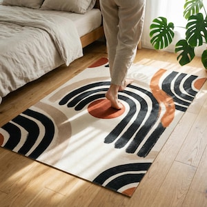 May include: A rectangular area rug with an abstract design. The rug features black, tan, and rust-colored curved lines and circles on an off-white background. The rug is placed on a hardwood floor near a bed.