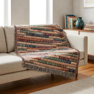 May include: A woven throw blanket featuring a horizontal striped pattern in shades of tan, brown, muted green, and deep blue. The blanket has a textured appearance with a fringed edge, draped over the back and seat of a light-colored sofa.