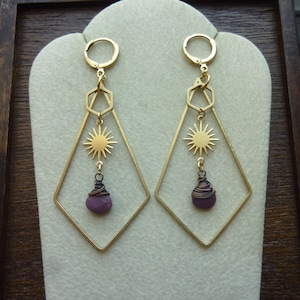 May include: A pair of gold-colored earrings with a geometric design. Each earring features a diamond-shaped frame, a sunburst charm, and a purple gemstone wrapped in wire. The earrings have a lever-back closure.