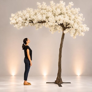 May include: A large artificial tree with a brown trunk and branches covered in white blossoms. The tree is on a metal base. A person wearing a black shirt and blue jeans is standing near the tree.