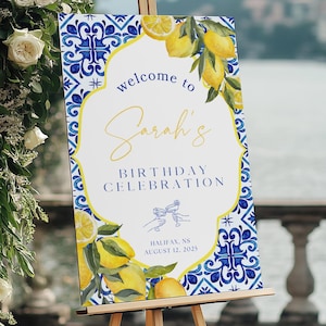 May include: A wooden easel displays a welcome sign for "Sarah's Birthday Celebration." The sign features a blue and white tile design with lemons and the text "Welcome to Sarah's Birthday Celebration, Halifax, NS August 12, 2023."