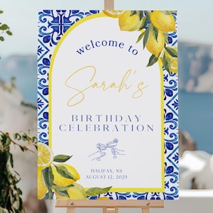 May include: A welcome sign for Sarah's birthday celebration, featuring a white background with blue and yellow accents. The sign includes lemons and a decorative border, displayed on a wooden easel. The text reads "Welcome to Sarah's Birthday Celebration."