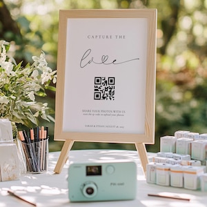 May include: A wooden framed sign with the text "Capture the love" and a QR code, displayed on a small easel. A mint green camera and stacks of film are on a white table, with pens and floral arrangements. The scene is set outdoors.