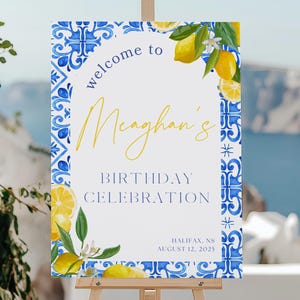 May include: A wooden easel displays a welcome sign for Meghan's birthday celebration. The sign features a white background with blue and yellow accents, including lemons and floral patterns. The text reads "Welcome to Meghan's Birthday Celebration" with the date and location.