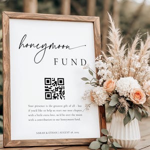 May include: A wooden-framed sign with the words "honeymoon fund" in script. It includes a QR code and text requesting contributions. A floral arrangement with peach and white roses is beside the sign. The sign is placed on a wooden surface, creating a romantic setting.