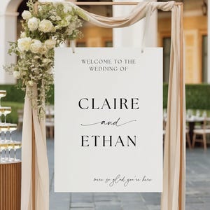 May include: A white wedding sign with the names "Claire and Ethan" in black lettering. The sign is part of a decorative arch with floral arrangements and draped fabric. The sign reads "Welcome to the Wedding of" and "were so glad you're here."