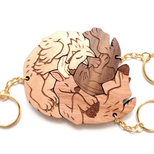 May include: Wooden puzzle keychain with a sleeping animal design. The keychain is made of layered wood in shades of brown and tan, with gold-coloured key rings and chains. The design is a puzzle of three animals.