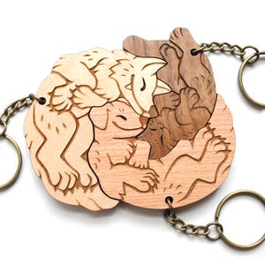 Peut inclure: Porte-clés en bois représentant trois animaux, un renard, un chien et un chat, finement sculptés et nichés ensemble. Le porte-clés est en bois brun clair et possède un anneau en métal doré.
