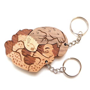 May include: Two wooden keyrings with a hedgehog design. The keyrings are made of light and dark brown wood, with a detailed illustration of a hedgehog hugging another hedgehog. Each keyring has a silver metal ring and chain.