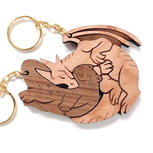 May include: Two wooden keychains featuring a dragon and otter design. The keychains are made of light and dark brown wood, with gold-colored key rings and chains. The dragon and otter are intertwined in a loving embrace. The keychains are on a white background.
