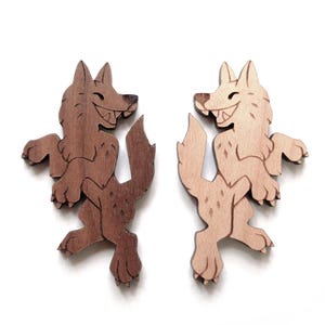May include: Two flat wooden wolf cutouts, one in a dark brown wood tone and one in a light tan wood tone. Each wolf is depicted in a standing, side-profile pose with a wide, toothy grin, pointed ears, and a bushy tail, featuring etched details.
