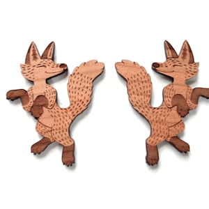 May include: A pair of wooden fox-shaped earrings featuring a light brown wood grain finish with darker brown engraved details on the fur, ears, and tail. Each fox is depicted in a standing pose with a bushy tail and closed eyes, facing inward.