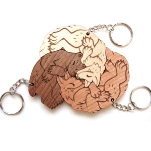May include: Wooden keyrings featuring a design of three intertwined animals in shades of brown and cream. Each keyring has a silver-coloured ring and chain. The keyrings are on a white background.