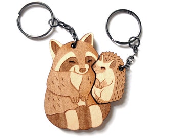 Puede incluir: Un llavero de madera con un mapache y un erizo abrazándose. El llavero está hecho de madera marrón claro y tiene un anillo de metal plateado.