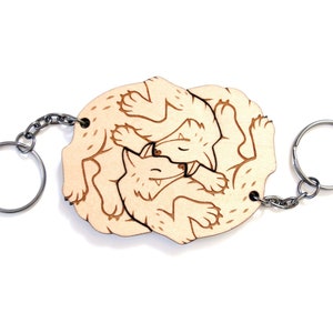 May include: Wooden keychain featuring a design of two wolves intertwined, creating a circular shape. The keychain is light brown and has a silver metal ring.