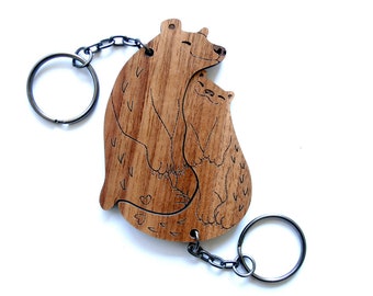 Könnte beinhalten: Holzschlüsselanhänger mit einem Bären und einer Katze, die sich umarmen. Der Schlüsselanhänger ist aus hellbraunem Holz und hat eine silberne Kette.