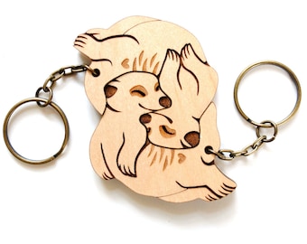 May include: Wooden keychain featuring two meerkats cuddling. The keychain is made of light brown wood and has a brass key ring.