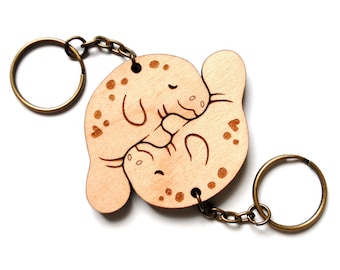 May include: A wooden keychain featuring two manatees hugging, with a brown chain and two metal rings. The manatees are carved in a light brown wood and have a natural wood finish.