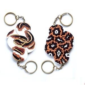 Könnte beinhalten: Zwei Schlüsselanhänger mit unterschiedlichen Designs. Einer zeigt eine braune und weiße Schlange, der andere ein braunes und schwarzes abstraktes Muster.