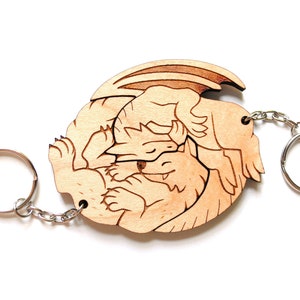 May include: A wooden keychain featuring a laser-cut design of two dragons curled up together. The keychain is attached to a silver metal ring.
