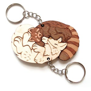 May include: Wooden keychain with a yin-yang design of two red pandas. One panda is light-coloured, the other is brown. The keychains are connected by a silver chain and ring. The background is white.