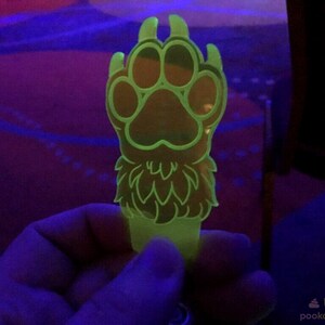 May include: A bright green glow-in-the-dark paw print keychain with a furry design. The paw print is detailed with a central pad and four surrounding toes.