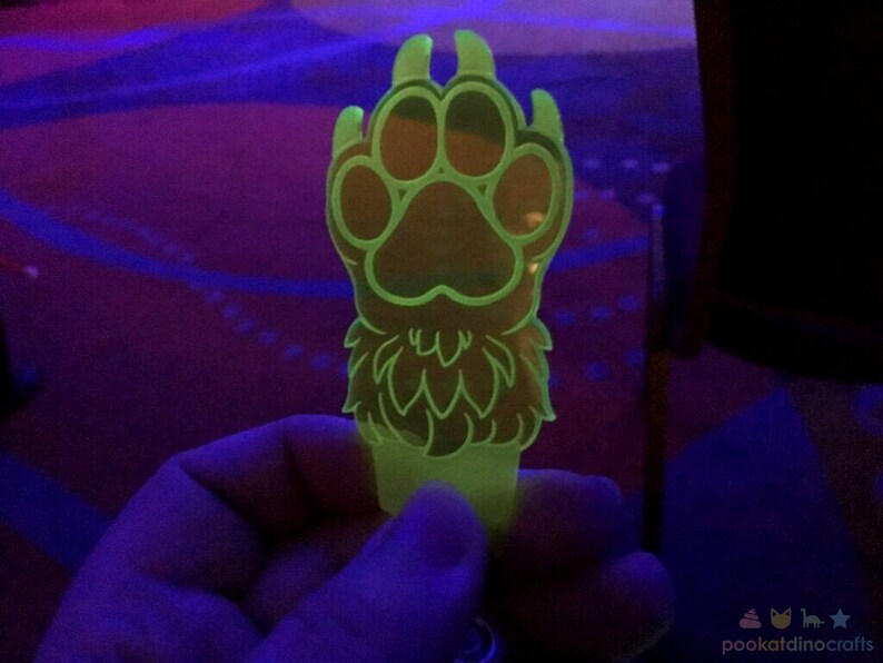 May include: A bright green glow-in-the-dark paw print keychain with a furry design. The paw print is detailed with a central pad and four surrounding toes.