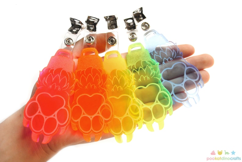 May include: A set of six colorful paw print keychains with a clear plastic strap and a metal clip. The paw prints are translucent and have a gradient effect, ranging from red to blue.