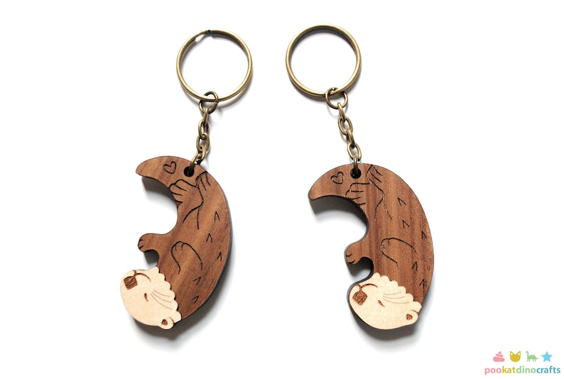 May include: Two wooden keyrings shaped like otters holding hearts. The otters are brown with white faces and are attached to gold key rings.