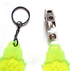May include: A lime green keychain with a floral design. The keychain has a metal ring and a clear plastic badge holder with a metal clip.