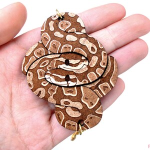 May include: A wooden snake keychain with a brown and white patterned design. The keychain is made of multiple pieces of wood that are layered and cut to create the snake shape. The keychain is attached to a gold-colored metal ring.