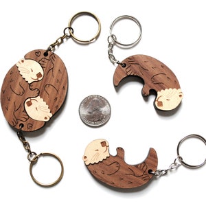 May include: Two wooden keyrings featuring a pair of otters holding each other. The keyrings are made of brown wood with a light brown otter design. The keyrings are attached to silver and bronze key rings.