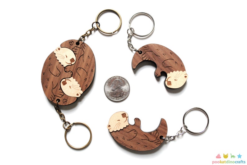 May include: Two wooden keyrings featuring a pair of otters holding each other. The keyrings are made of brown wood with a light brown otter design. The keyrings are attached to silver and bronze key rings.
