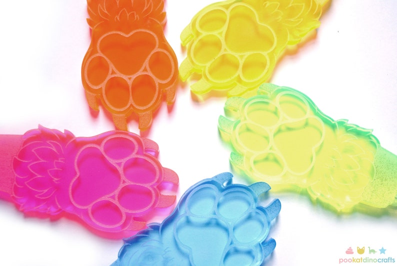 May include: Five colorful paw print keychains made of clear plastic. The keychains are in shades of orange, yellow, green, pink, and blue. Each keychain has a paw print design with three toes and a small, stylized tail.