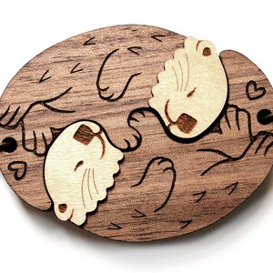 May include: A wooden keychain featuring two otters holding paws and sleeping. The otters are carved in a light brown colour and the background is a darker brown. The keychain has a metal chain attached.