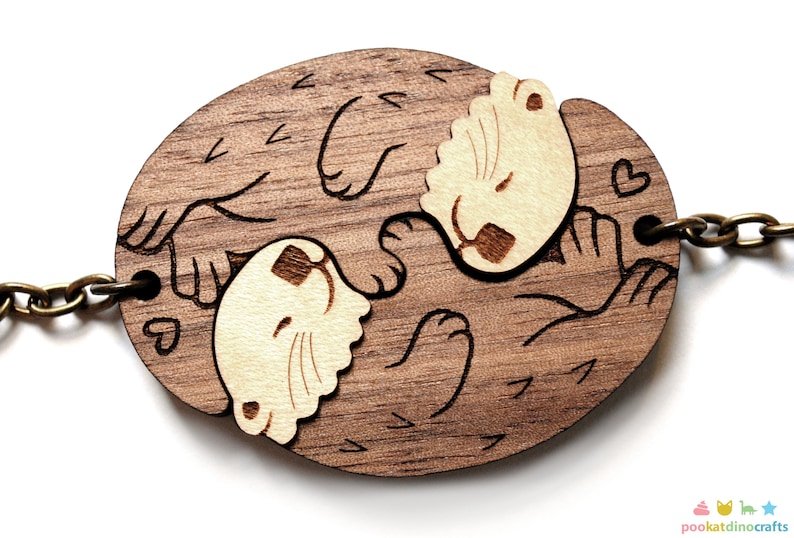 May include: A wooden keychain featuring two otters holding paws and sleeping. The otters are carved in a light brown colour and the background is a darker brown. The keychain has a metal chain attached.