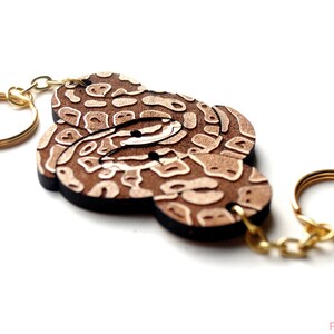 May include: A wooden keychain featuring a brown and white patterned design, resembling a snake skin. The keychain has a gold metal ring and chain.