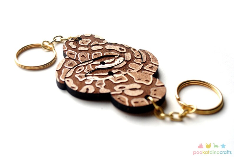 May include: A wooden keychain featuring a brown and white patterned design, resembling a snake skin. The keychain has a gold metal ring and chain.