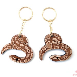 May include: Two brown wooden snake keychains with gold key rings. The snakes are curled up in a circle and have a brown and white pattern.