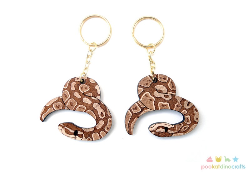 May include: Two brown wooden snake keychains with gold key rings. The snakes are curled up in a circle and have a brown and white pattern.