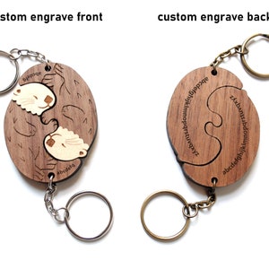 May include: A wooden keychain featuring a pair of otters hugging. The keychain is split into two pieces that fit together like a puzzle. The back of the keychain has the alphabet engraved on it.