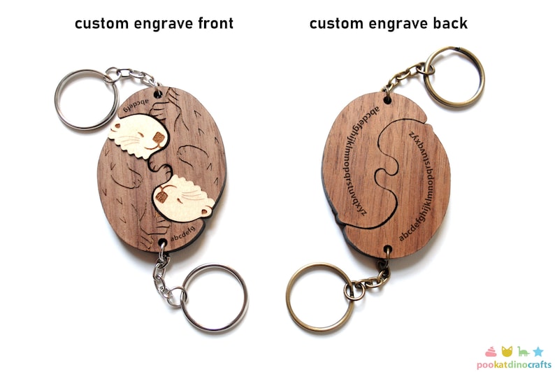 May include: A wooden keychain featuring a pair of otters hugging. The keychain is split into two pieces that fit together like a puzzle. The back of the keychain has the alphabet engraved on it.
