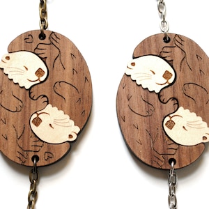 May include: Two wooden keyrings featuring a design of two otters cuddling. The otters are carved in a light brown colour and the background is a darker brown. The keyrings are attached to silver and gold chains.