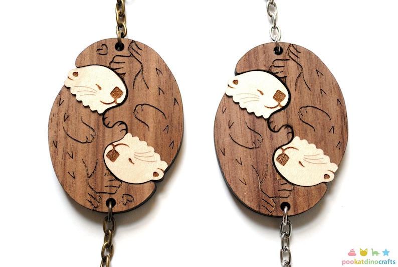 May include: Two wooden keyrings featuring a design of two otters cuddling. The otters are carved in a light brown colour and the background is a darker brown. The keyrings are attached to silver and gold chains.