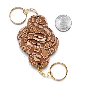 May include: A wooden keychain featuring a brown and white snake design. The snake is coiled and has a realistic pattern. The keychain is attached to a gold chain and ring.