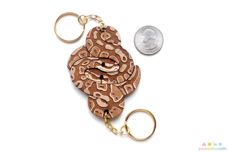 May include: A wooden keychain featuring a brown and white snake design. The snake is coiled and has a realistic pattern. The keychain is attached to a gold chain and ring.