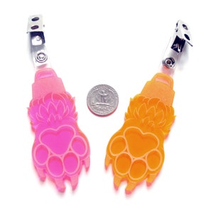 May include: Two colorful paw print shaped badge holders. The left holder is pink and the right holder is orange. Both holders have a silver clip on the top.