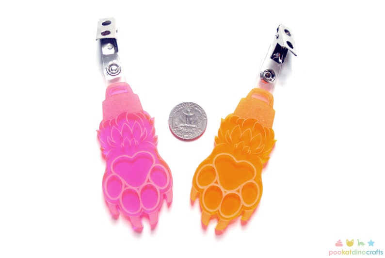 May include: Two colorful paw print shaped badge holders. The left holder is pink and the right holder is orange. Both holders have a silver clip on the top.
