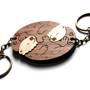 May include: Wooden keychain featuring two otters cuddling, carved in a light brown colour with a darker brown outline. The otters are holding hearts and the keychain has a bronze metal ring.